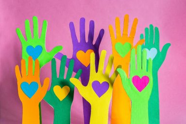 Multi-colored paper hands against a pink background. They all have different colored hearts on their palms.